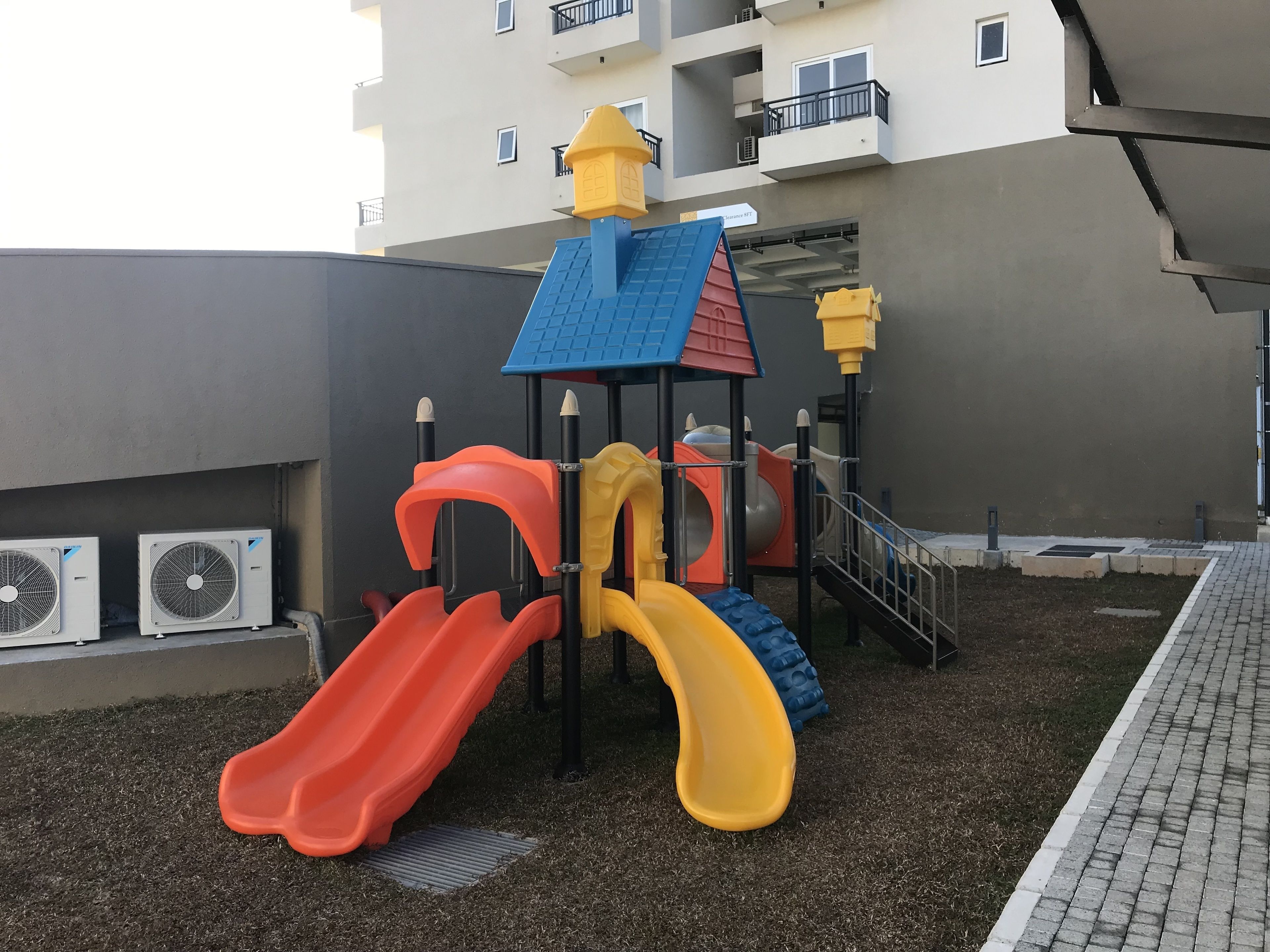 A playground with slides and climbing structures, surrounded by a building with balconies and air conditioning units.