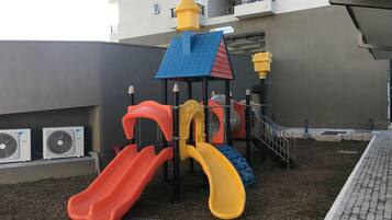A playground with slides and climbing structures, surrounded by a building with balconies and air conditioning units.