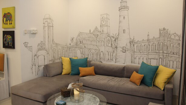 A living room with a sofa, cushions, a glass coffee table, and a wall mural of a cityscape.