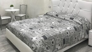 Un dormitorio con cama blanca, colcha estampada, una mesita redonda con dos sillas y un cuadro con corazones.