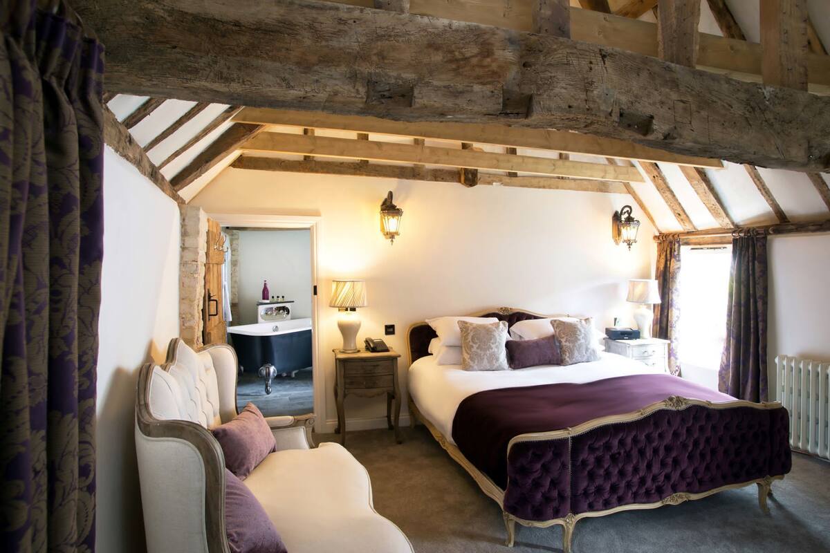 A bedroom with a large bed, a bedside table, a chair, and a bathroom visible through an open door.