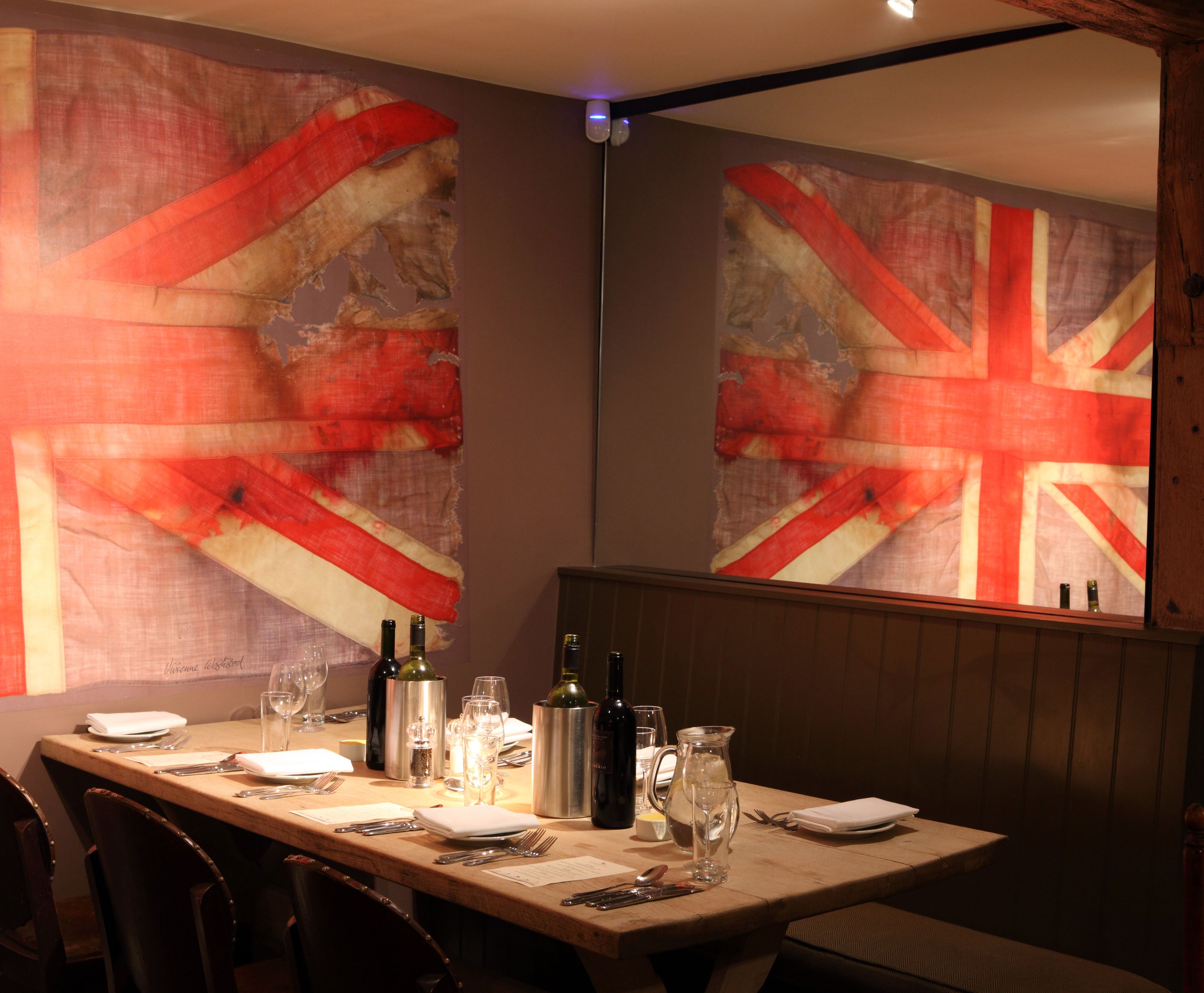 Mesa de jantar posta com garrafas de vinho, copos e talheres, com um grande mural na parede com a Union Jack.