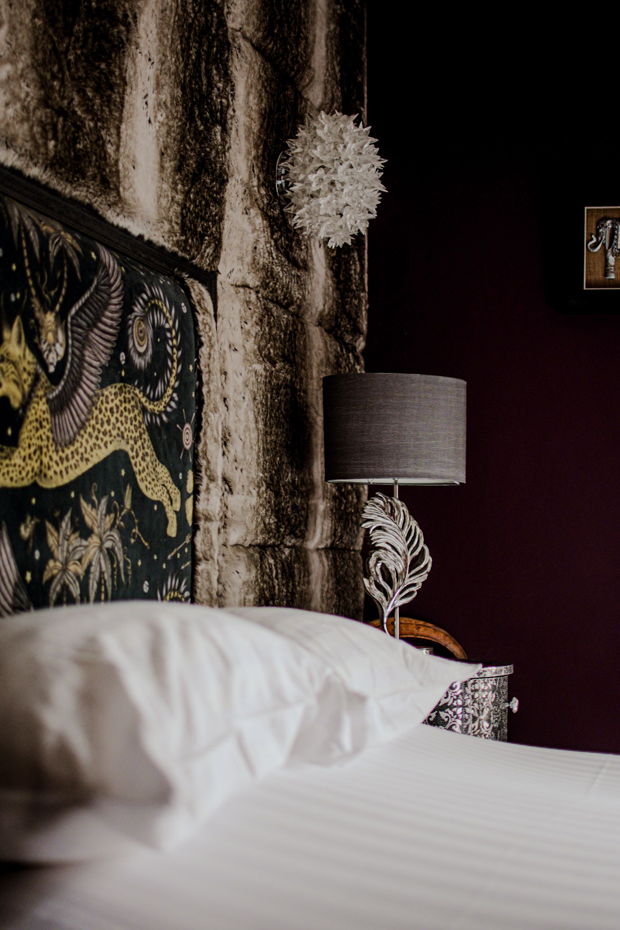 A bed with white linens, a decorative tapestry with a leopard design, a dark wall, and a lamp with a feather design.