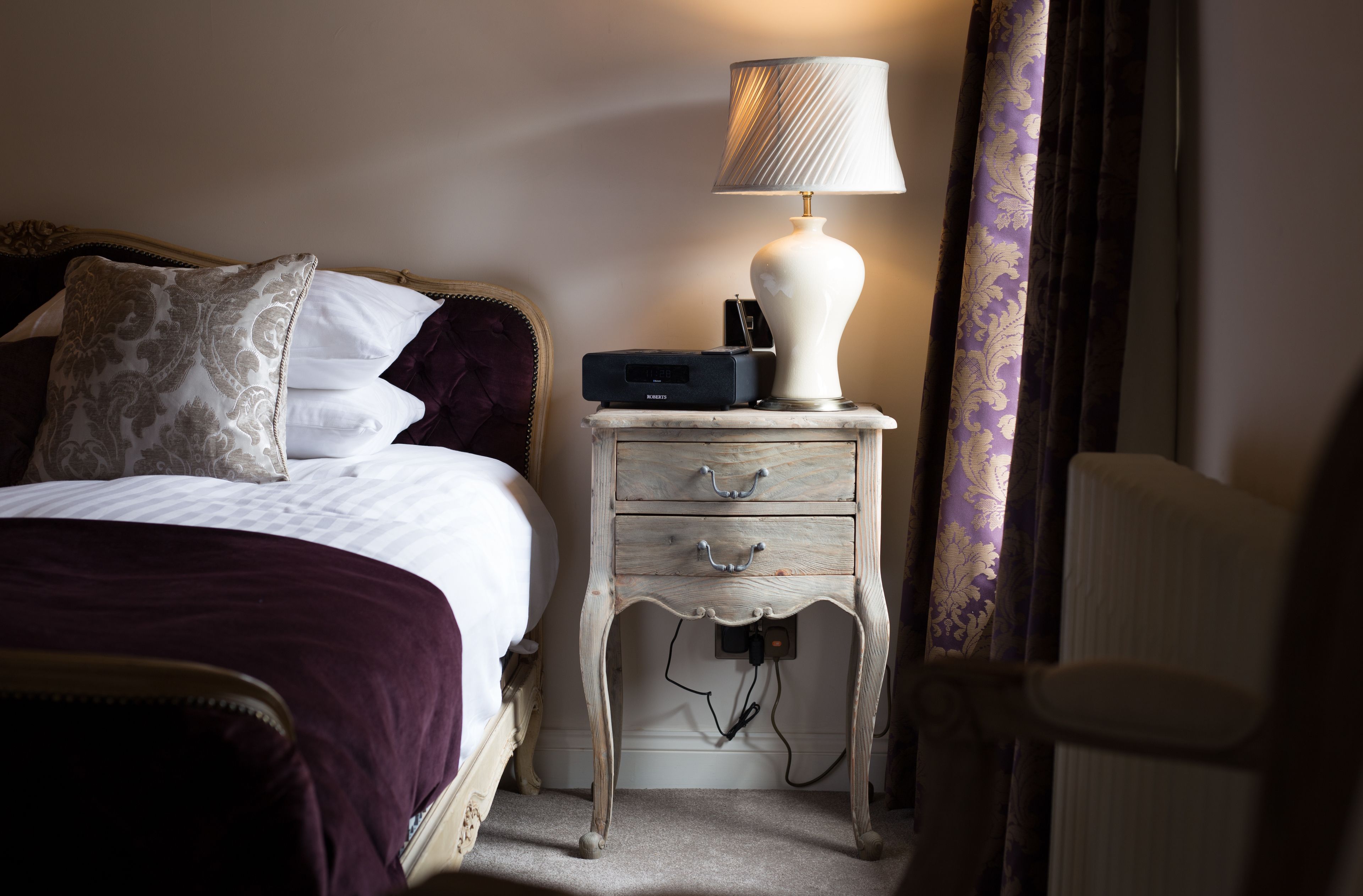 A neatly made bed with a purple blanket, a white lamp on a wooden nightstand, and a power outlet.