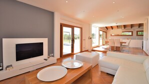 A modern living room with a flat-screen TV, a dining area with a table and chairs, and a kitchen in the background.