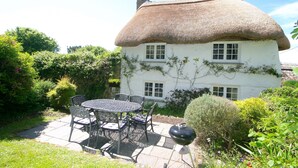 A white house with a thatched roof, a patio with a round table and chairs, and a barbecue grill. - Putsborough Manor Cottage (4 + Cot) Dogs Tennis Beach Jacuzzi Smart TV wifi (Croyde)