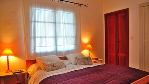 A neatly made bed with a purple bedspread, two bedside lamps, and a window with sheer curtains.