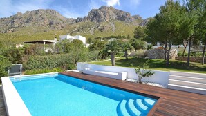 A swimming pool with wooden deck, surrounded by greenery and white walls, with mountains in the background.