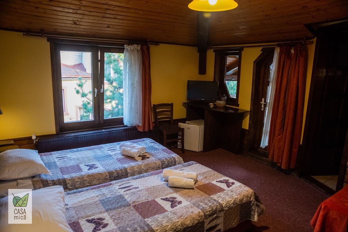 A room with two beds, a desk, a small refrigerator, and a window with curtains.