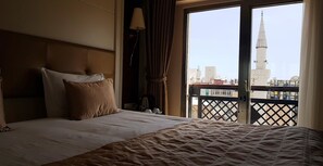 Une chambre d’hôtel avec un grand lit, une tête de lit, une lampe et une vue sur le paysage urbain, avec une tour visible depuis le balcon.