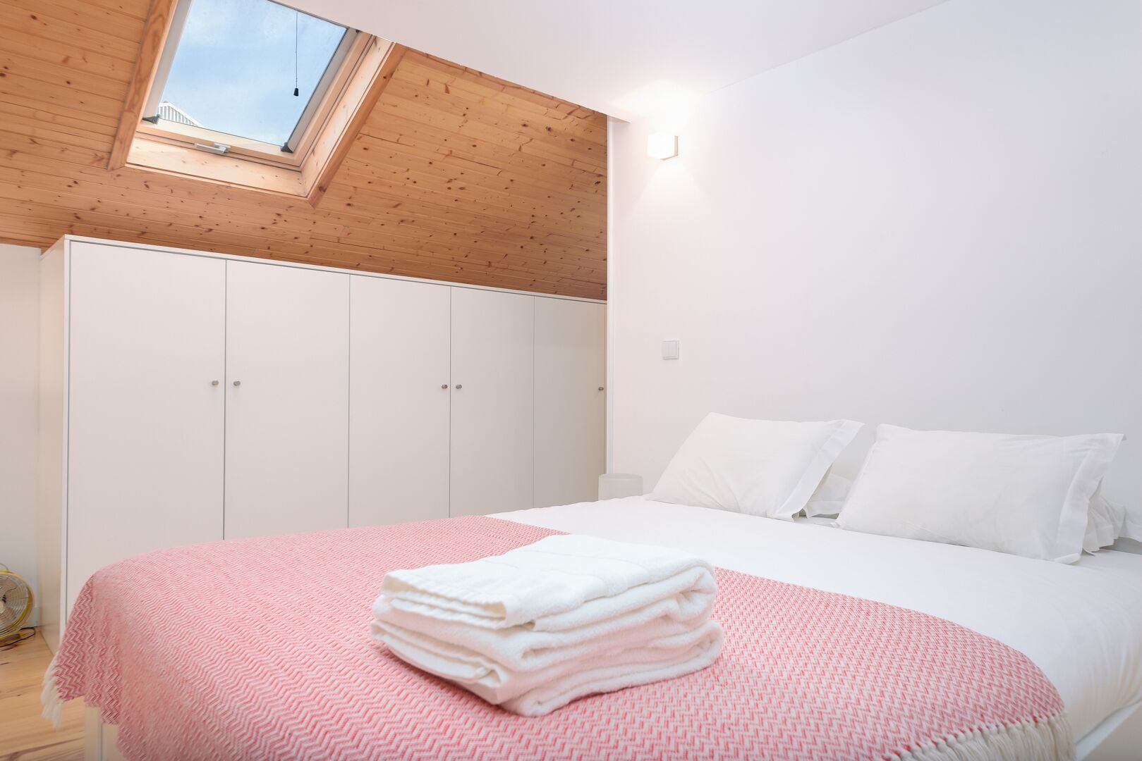 Un dormitorio con tragaluz, armario blanco y cama con ropa de cama blanca y una manta rosa.