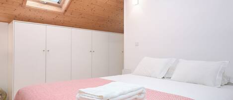 Un dormitorio con tragaluz, armario blanco y cama con ropa de cama blanca y una manta rosa.