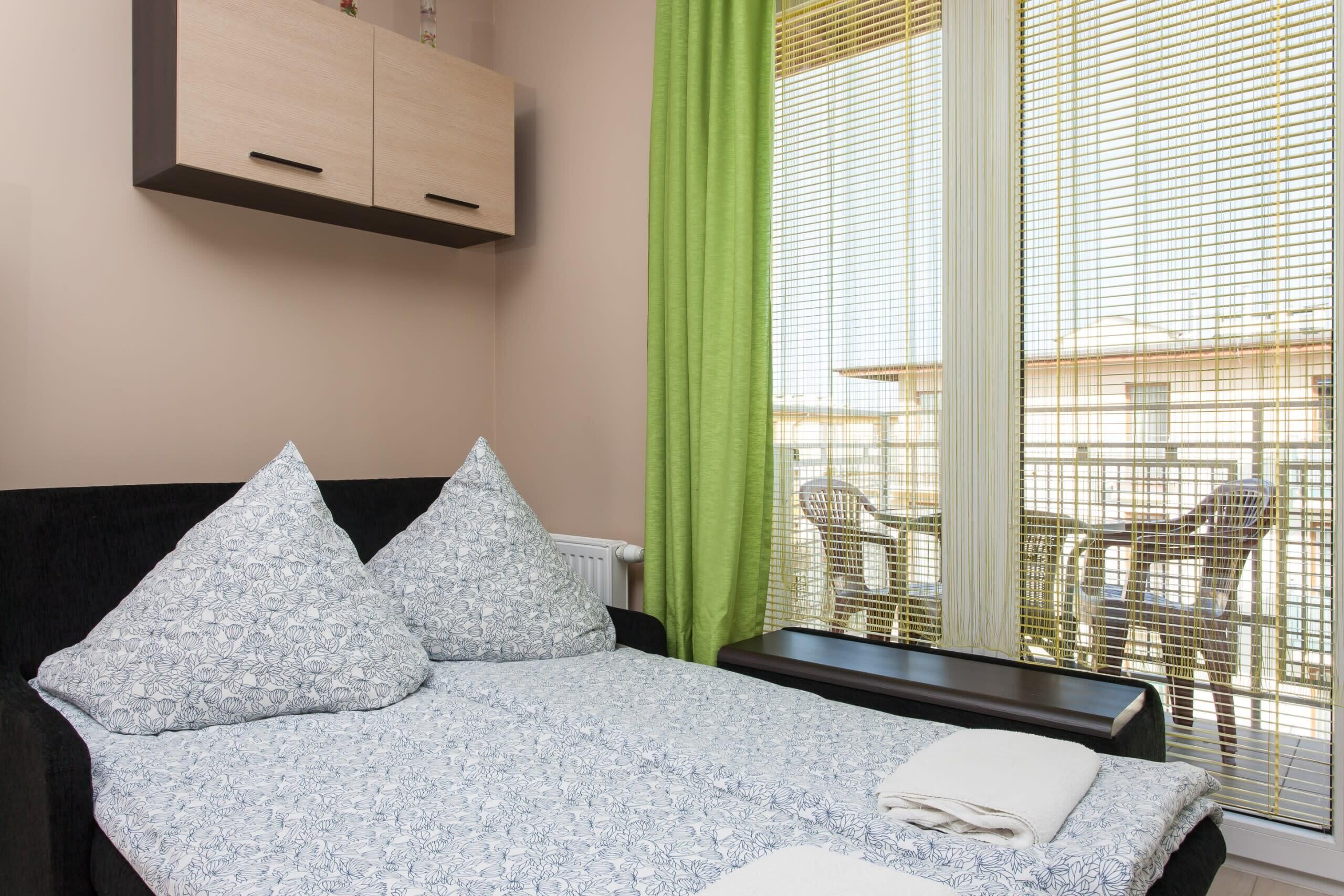 Un dormitorio con cama, almohadas, mesita de noche, ventana con persianas y vistas a muebles de exterior.