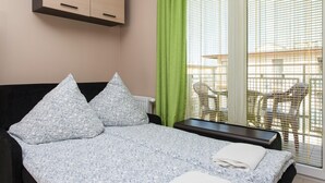 A bedroom with a bed, pillows, a bedside table, a window with blinds, and a view of outdoor furniture.