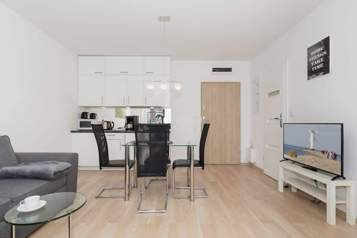 Un salon moderne avec un canapé gris, une table basse en verre, un coin repas avec une table et des chaises en verre, une cuisine avec des meubles blancs, une porte en bois et un téléviseur posé sur un meuble blanc.