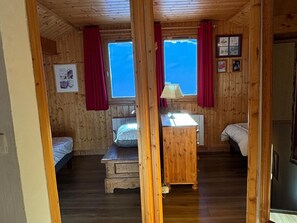 Une chambre dans un chalet en bois, avec deux lits, une petite table et une fenêtre ornée de rideaux rouges.