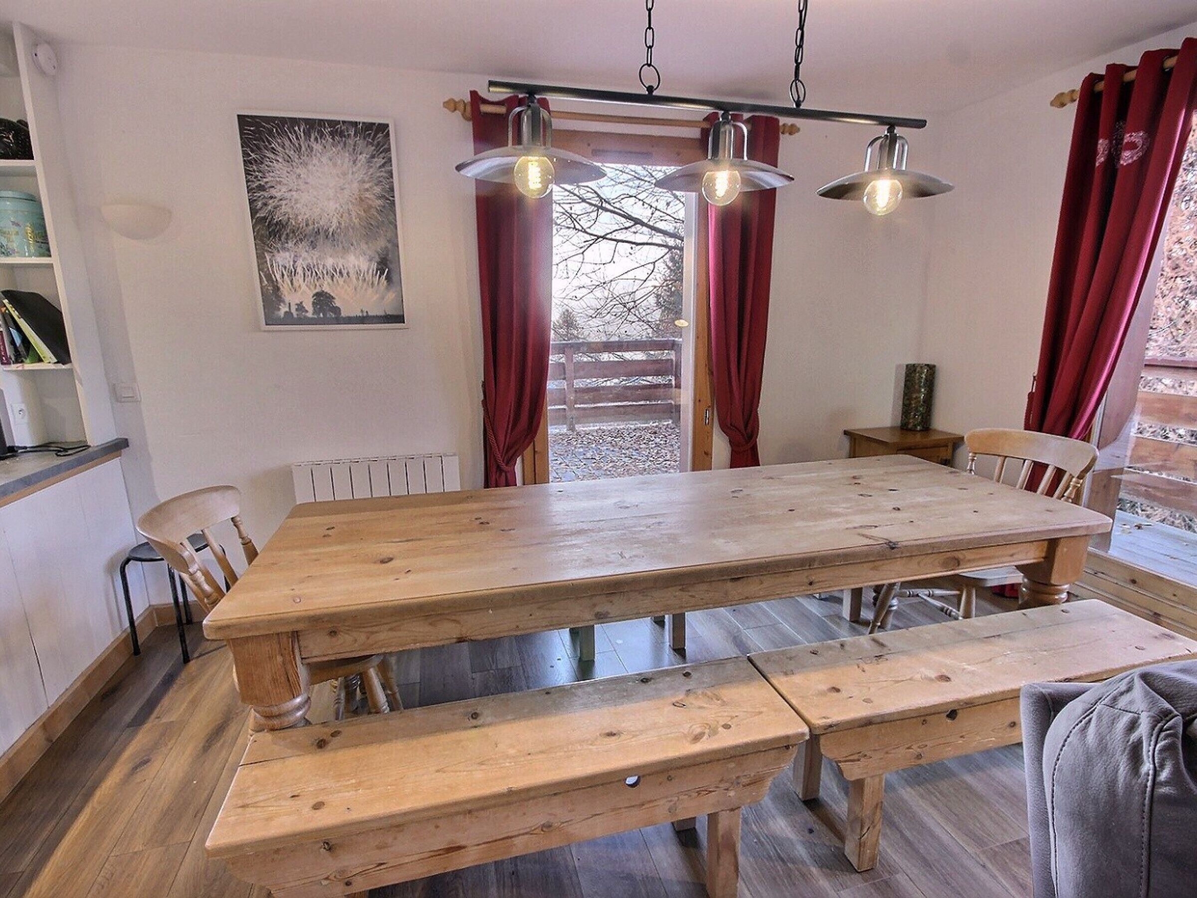 Un espace repas avec une table et des bancs en bois, des luminaires suspendus et une vue sur une terrasse en bois, visible à travers une fenêtre.