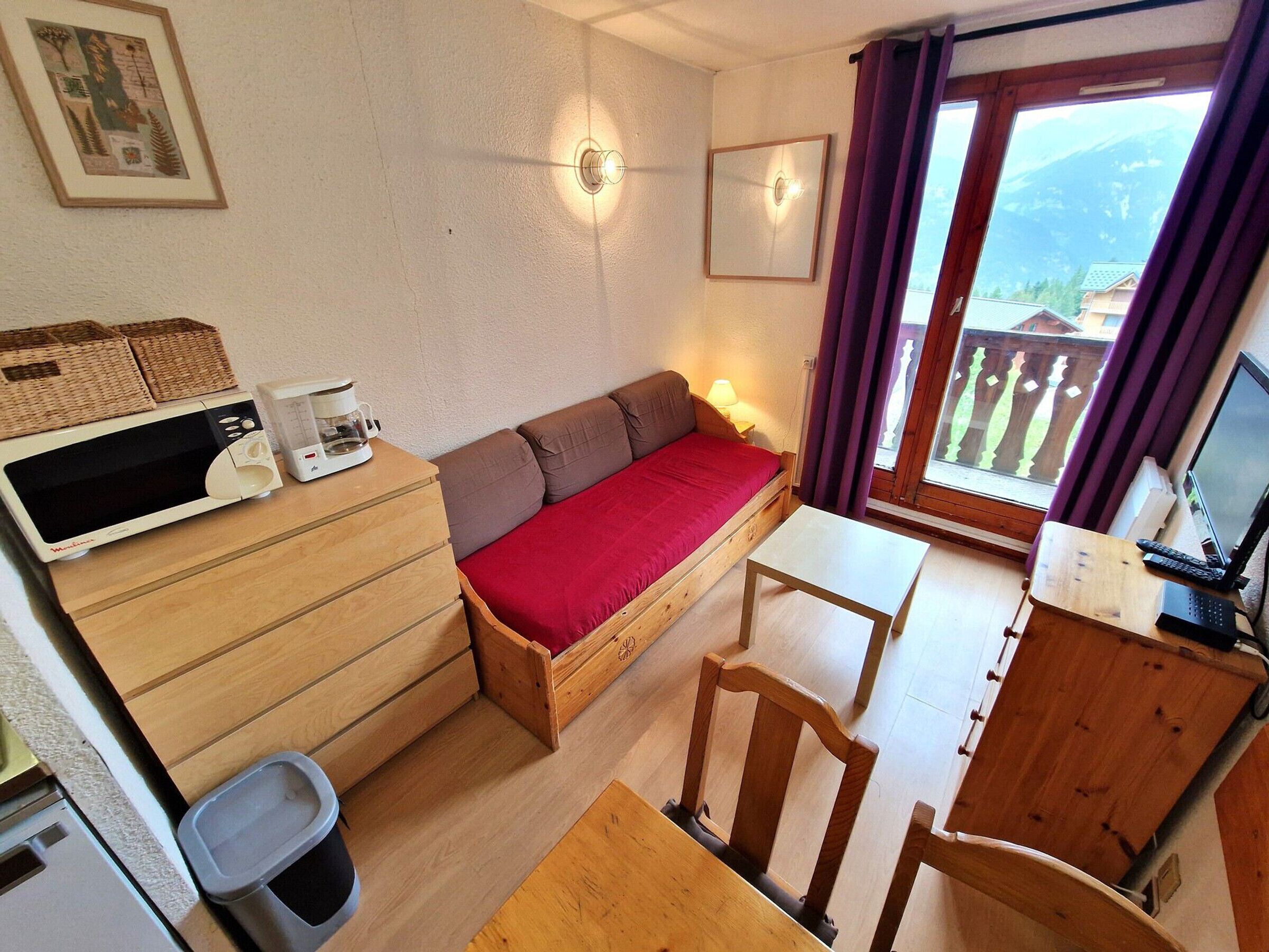A compact room with a bed, a small desk, a chair, a microwave, a trash bin, and a window with a view of mountains.