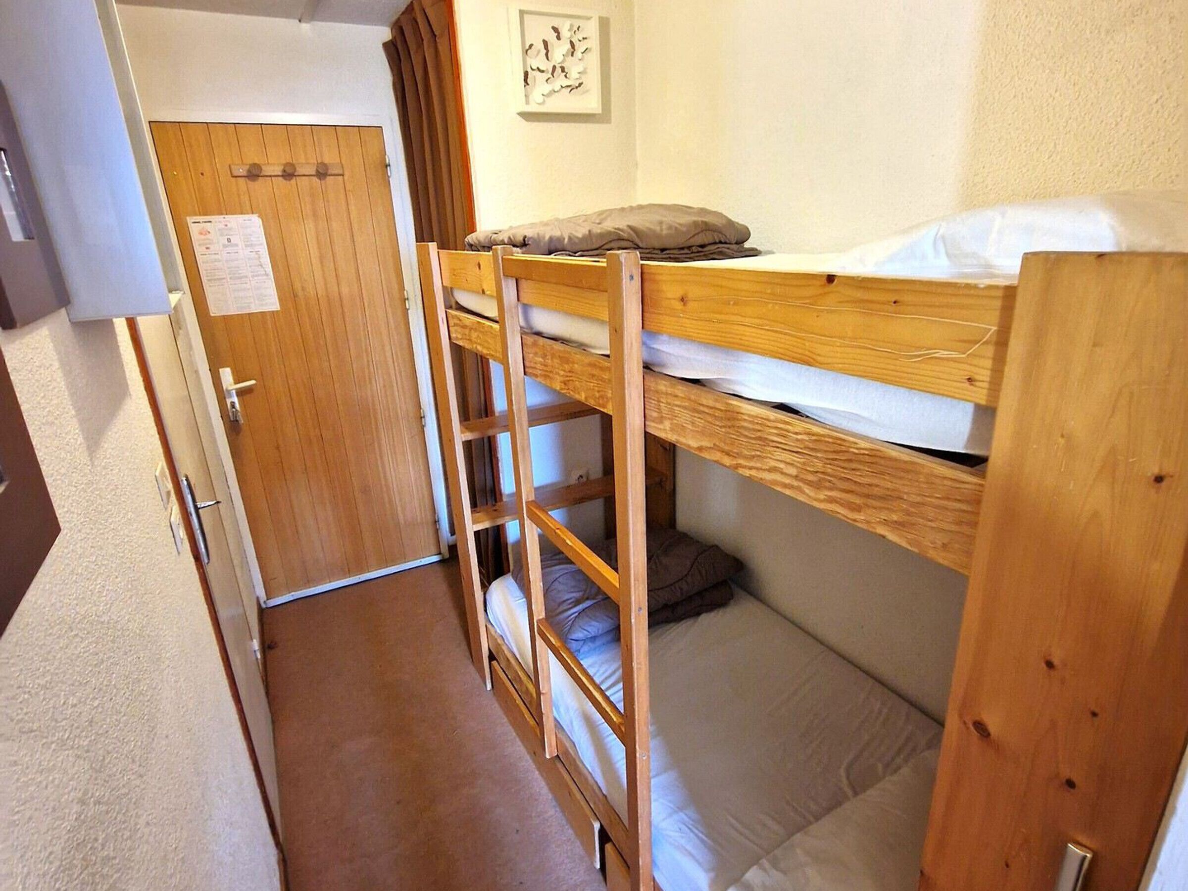 A narrow room with bunk beds, a wooden door, and a small white wall with a framed picture.