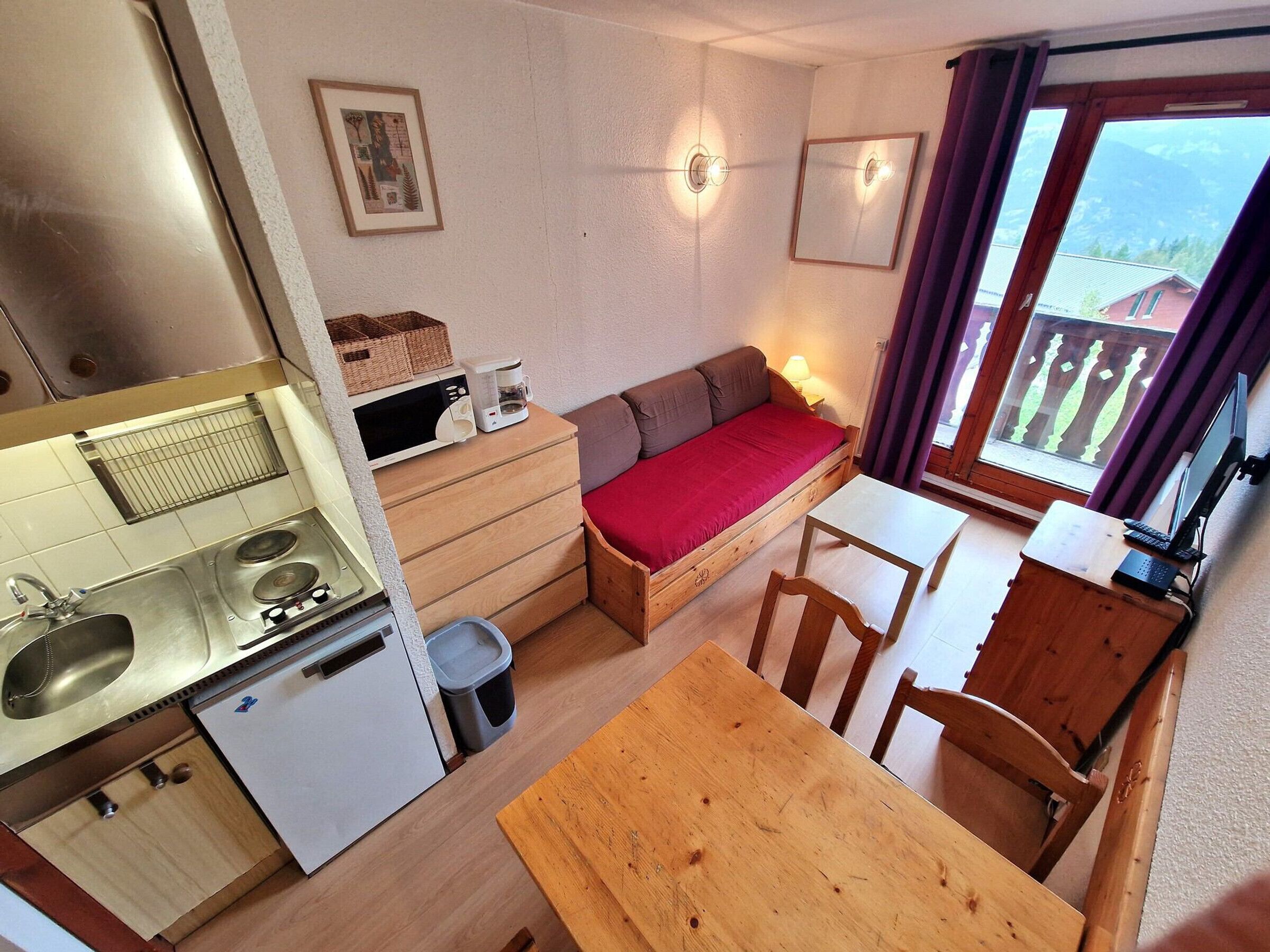 A compact kitchen with a sink, stove, and refrigerator. There is a wooden table with chairs, a small sofa with a red cushion, and a window with curtains.