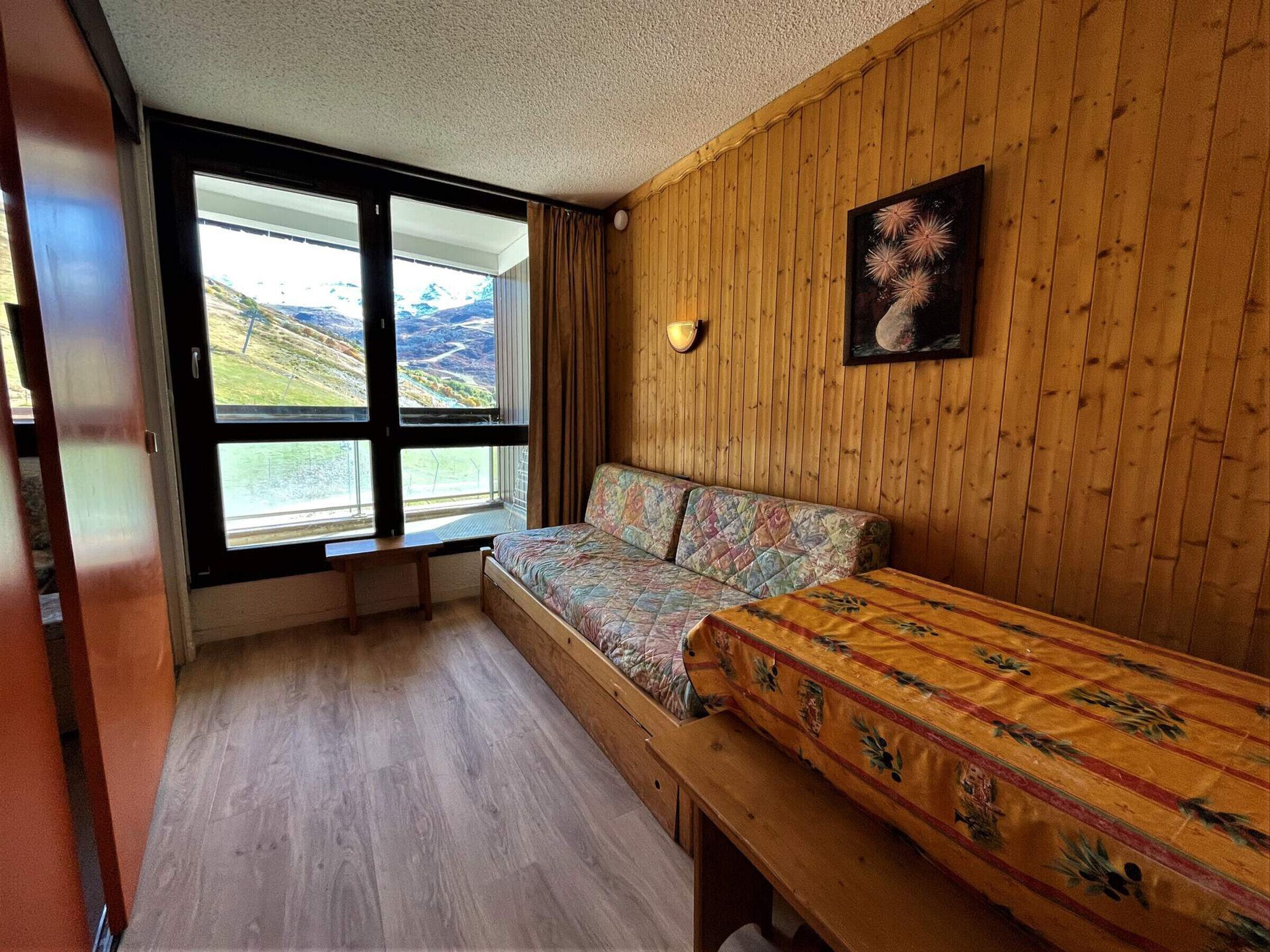 A room with a large window, wooden walls, a bed with a colorful quilt, a bench, and a wall-mounted light fixture.