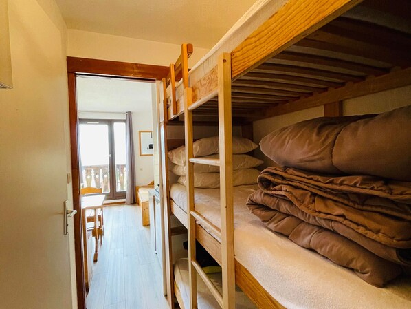 A narrow hallway with a wooden bunk bed on the right, a door leading to a balcony, and a small table with chairs in the background.