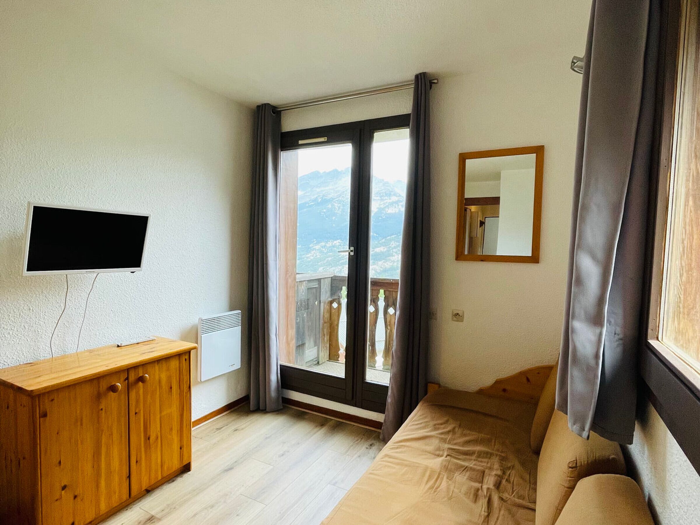 A room with a wooden cabinet, a flat-screen TV, a bed, a window with a view of mountains, and a mirror on the wall.