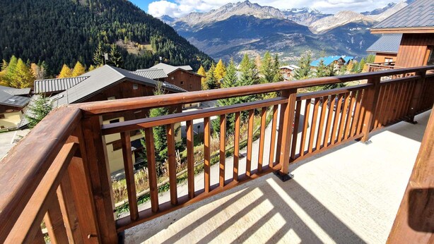 A wooden balcony with a view of a mountainous landscape, including houses and a clear sky.