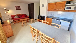 A compact kitchen with a microwave, stove, and sink. There's a wooden dining table with chairs and a red sofa in the living area.