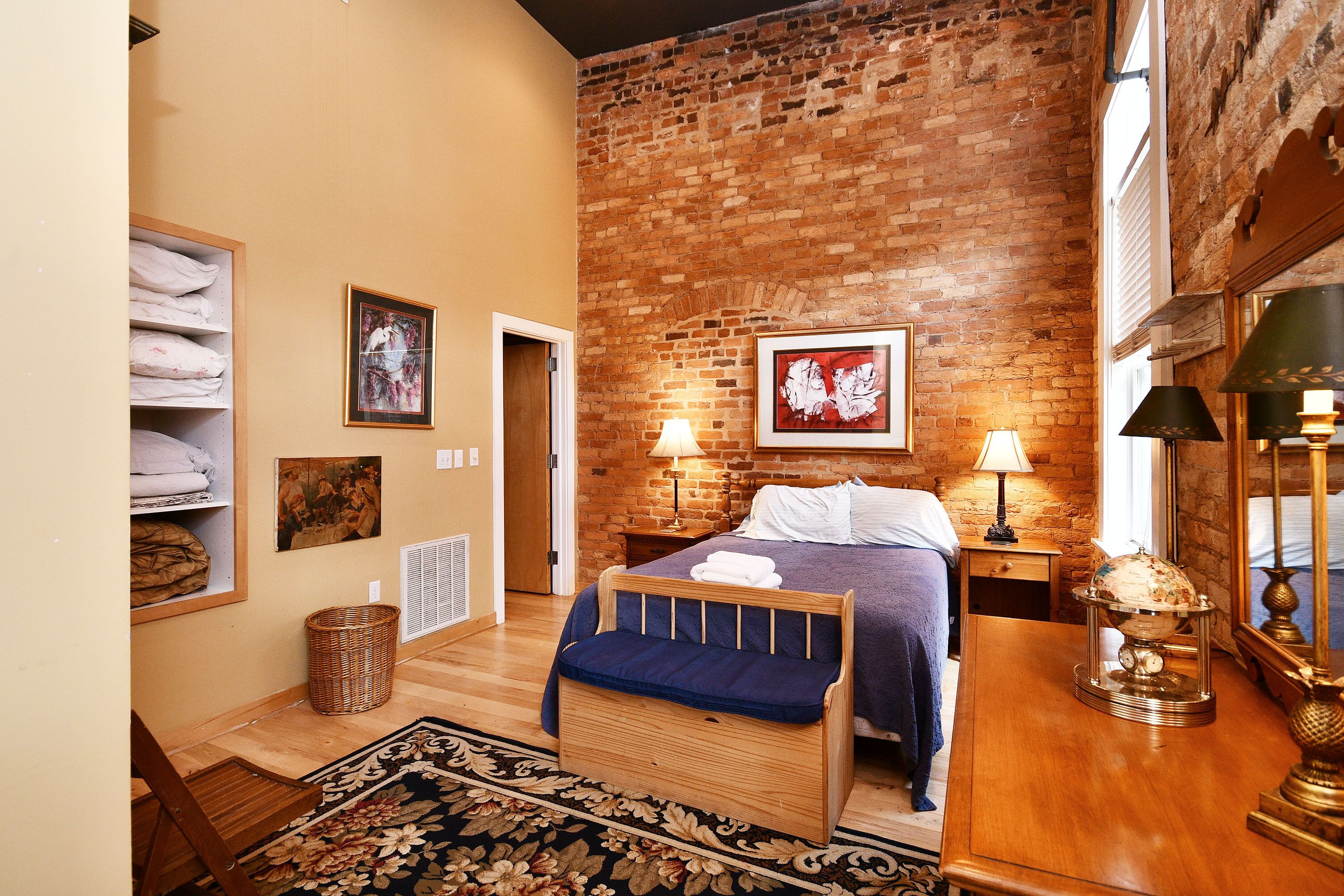 A bedroom with a brick wall, a bed with a blue blanket, a wooden nightstand with a lamp, and a globe on the nightstand.