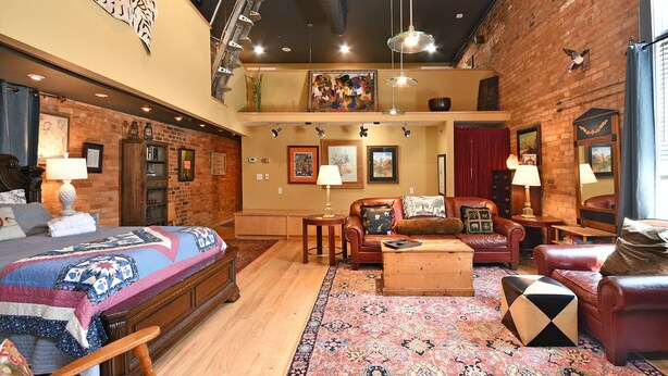 A spacious loft-style room with a large bed, multiple seating areas, and a high ceiling with exposed brick walls. - Luxury Loft and Rooftop NOMA Square, Sleeps 8-10 (Greenville)