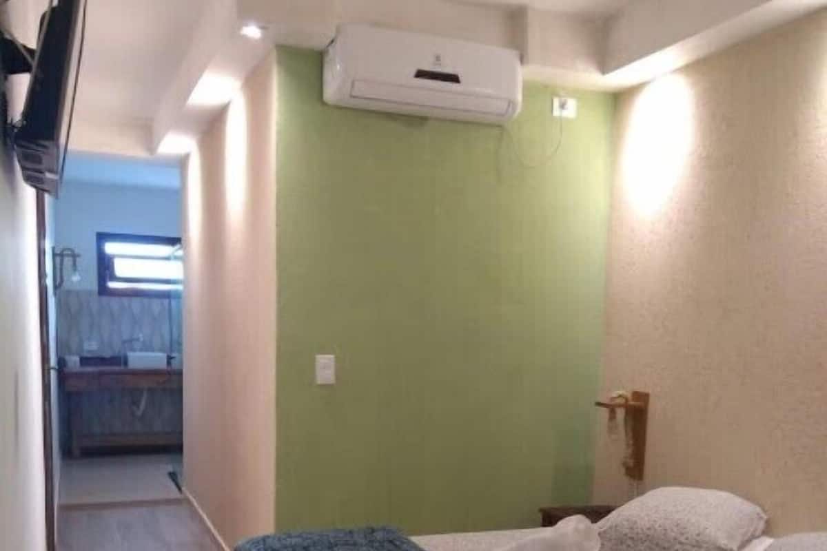 A bedroom with a bed, a green wall, an air conditioning unit, and a hallway leading to a bathroom.