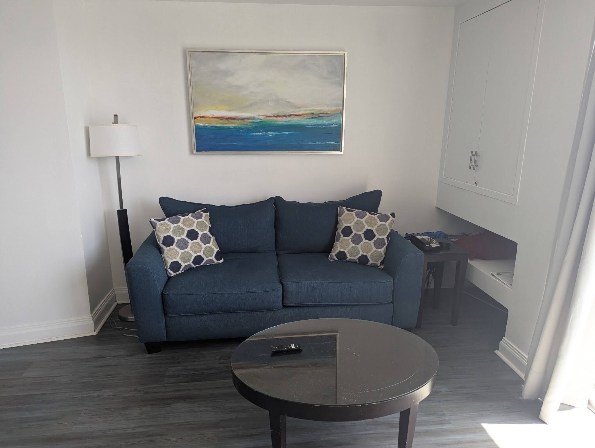 Ett vardagsrum med en blå soffa, två mönstrade kuddar, ett runt svart soffbord, en golvlampa och en tavla som föreställer ett landskap.