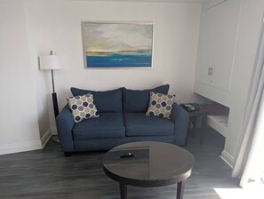 A living room with a blue sofa, two patterned pillows, a round black coffee table, a floor lamp, and a painting of a landscape.