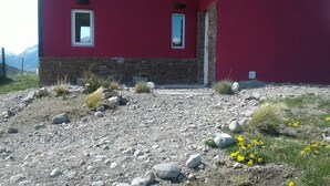 Um edifício vermelho com base de pedra, duas janelas e uma entrada com calçamento de pedras e flores amarelas.