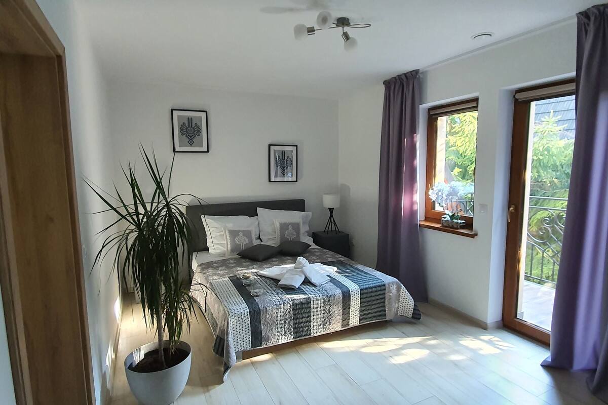 A bedroom with a bed, a potted plant, a window with curtains, and framed pictures on the wall.