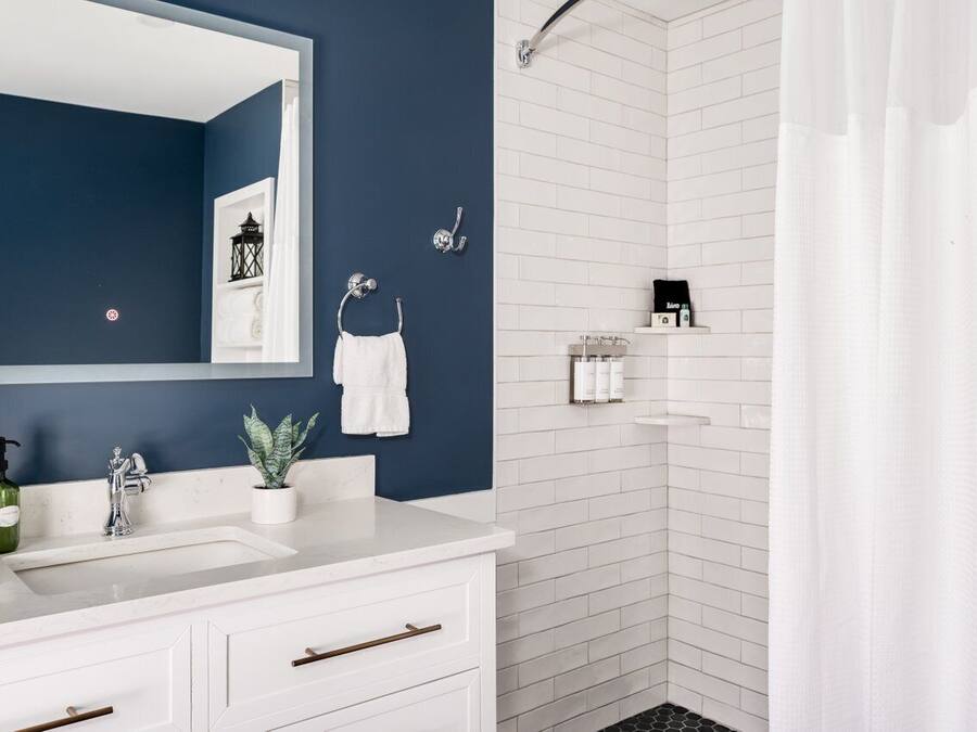 Uma casa de banho moderna com uma parede azul escuro, azulejos brancos, um espelho grande, um lavatório com um frasco verde e zona de duche.