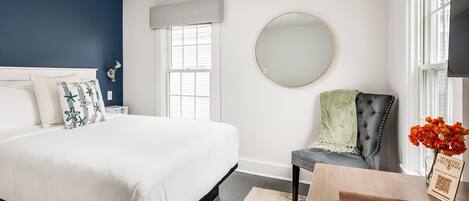 A bedroom with a bed, a round mirror, a chair, and a table with a vase of flowers.
