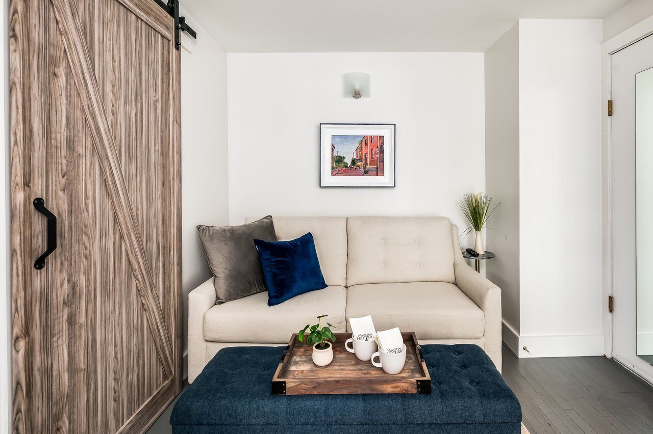 En hyggelig stue med en beige sofa, en trædør, et billede i ramme på væggen, og en bakke med krus samt en plante på gulvet.
