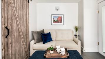 En hyggelig stue med en beige sofa, en trædør, et billede i ramme på væggen, og en bakke med krus samt en plante på gulvet.