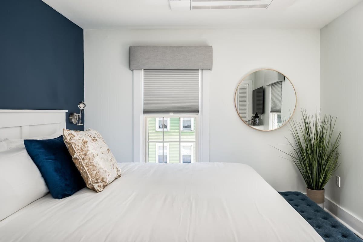 A bedroom with a bed, a round mirror, a window with blinds, and a potted plant.