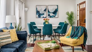 A modern living room with a dark blue sofa, teal chairs, a yellow armchair, a coffee table, and a dining table with chairs. There are two abstract paintings on the wall, a potted plant, and a door in the background.