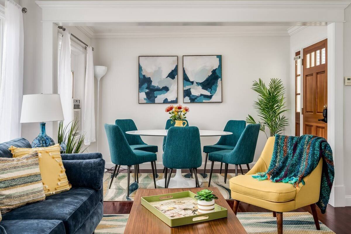 A modern living room with a dark blue sofa, teal chairs, a yellow armchair, a coffee table, and a dining table with chairs. There are two abstract paintings on the wall, a potted plant, and a door in the background.