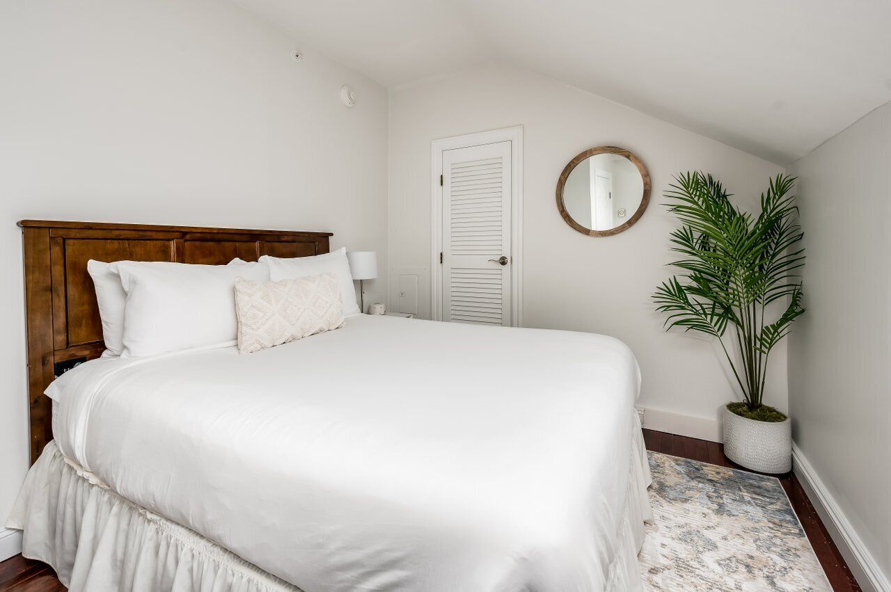A bedroom with a wooden headboard, white bedding, a round mirror, a potted plant, and a white door.
