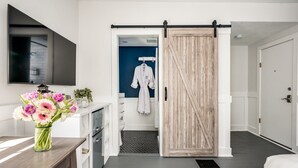 A modern hotel room with a sliding door leading to a bathroom, a flat-screen TV mounted on the wall, and a vase of flowers on the dresser.