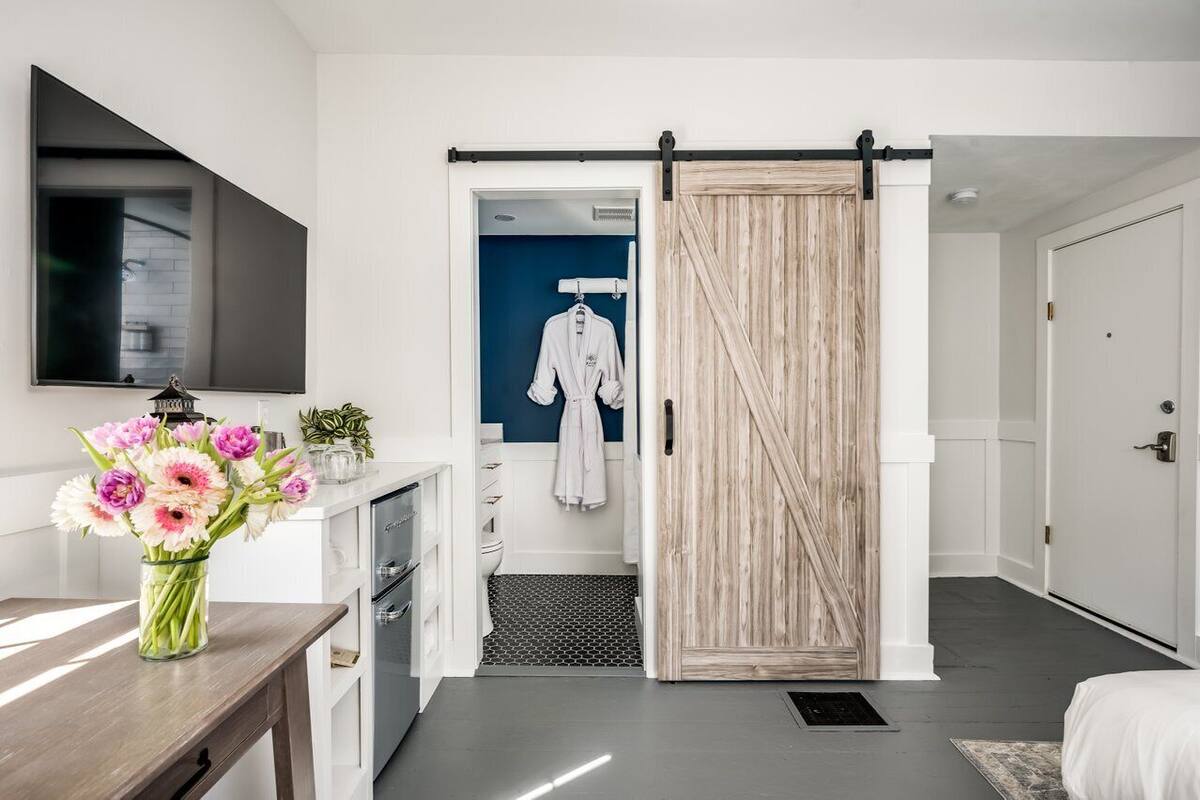A modern hotel room with a sliding door leading to a bathroom, a flat-screen TV mounted on the wall, and a vase of flowers on the dresser.