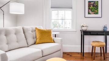 A modern living room with a grey sofa, a mustard-colored cushion, a small round ottoman, a desk with a chair, and a framed painting on the wall.