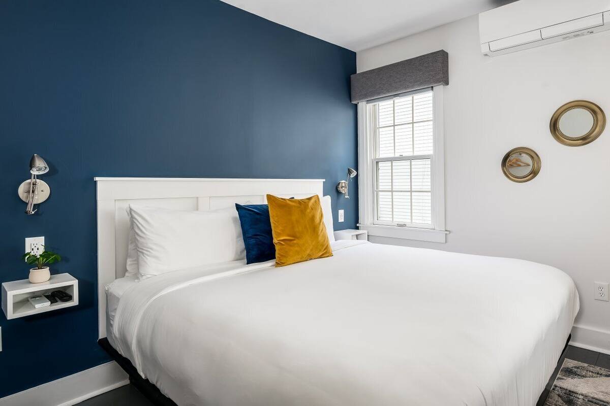 A bedroom with a white bed, blue walls, a window with blinds, a bedside table with a lamp, and a small shelf with a plant.