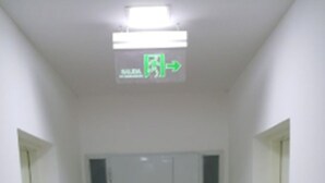 Un pasillo estrecho con una puerta blanca al final, un letrero de salida verde arriba y un suelo de baldosas.