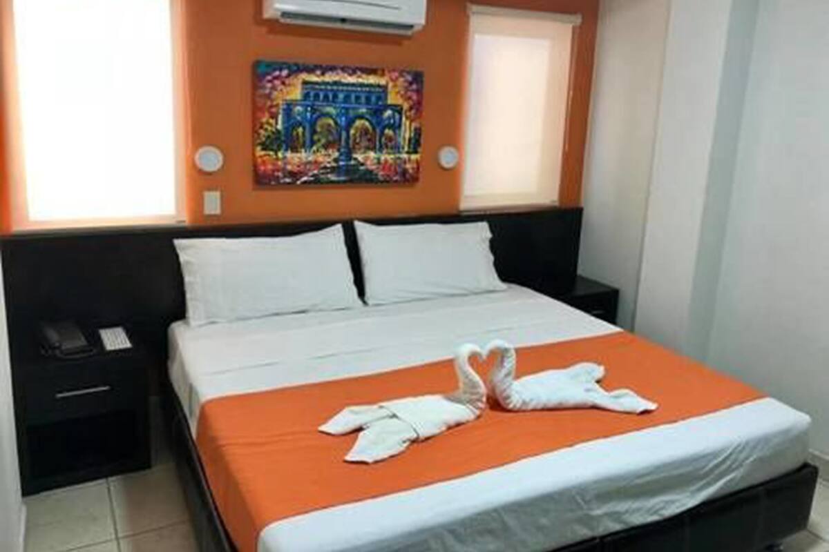 A hotel room with a bed, an air conditioner, a painting, and a window.